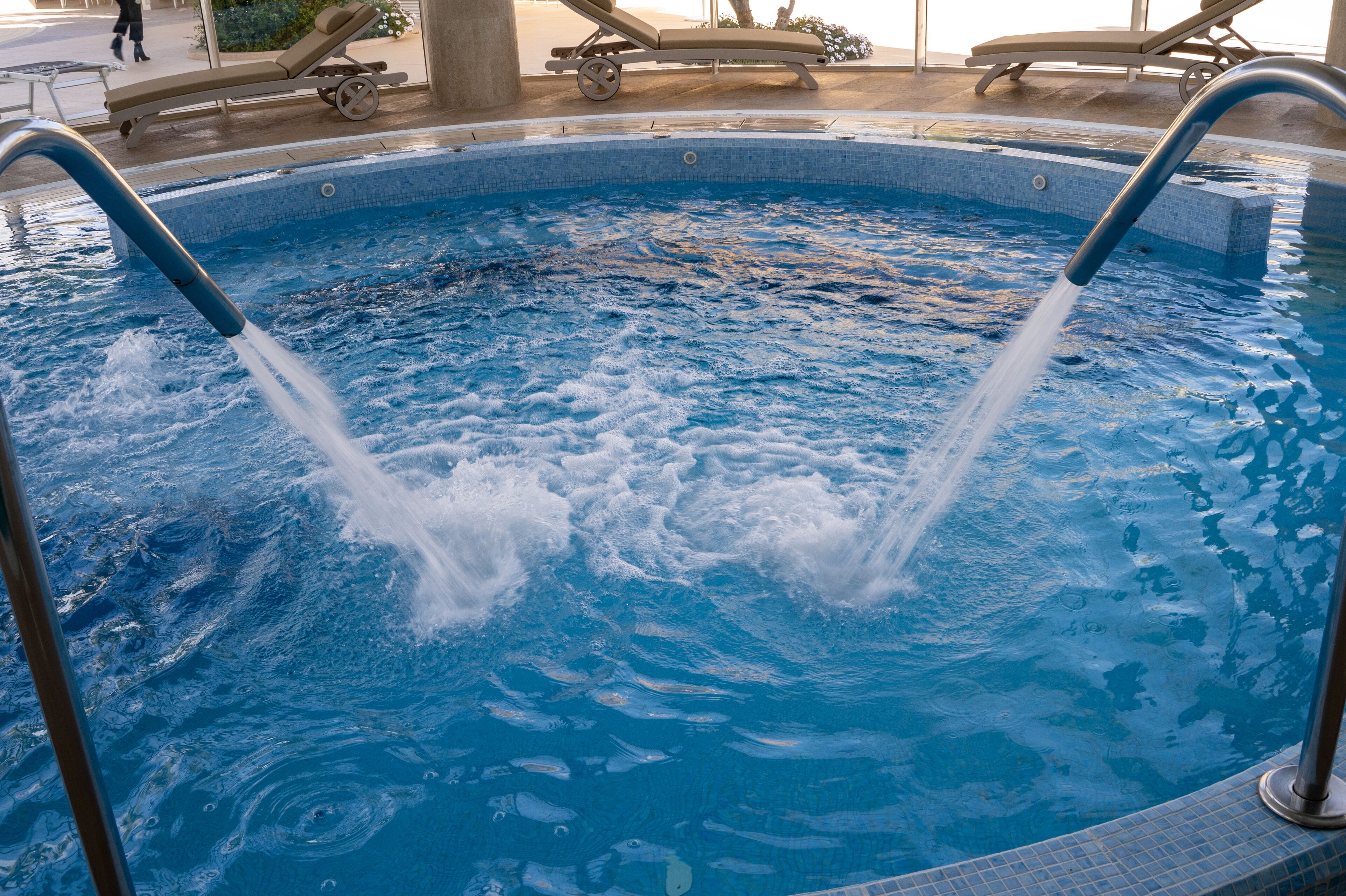 A swimming pool with two water jets, lounge chairs, and a tiled deck area.