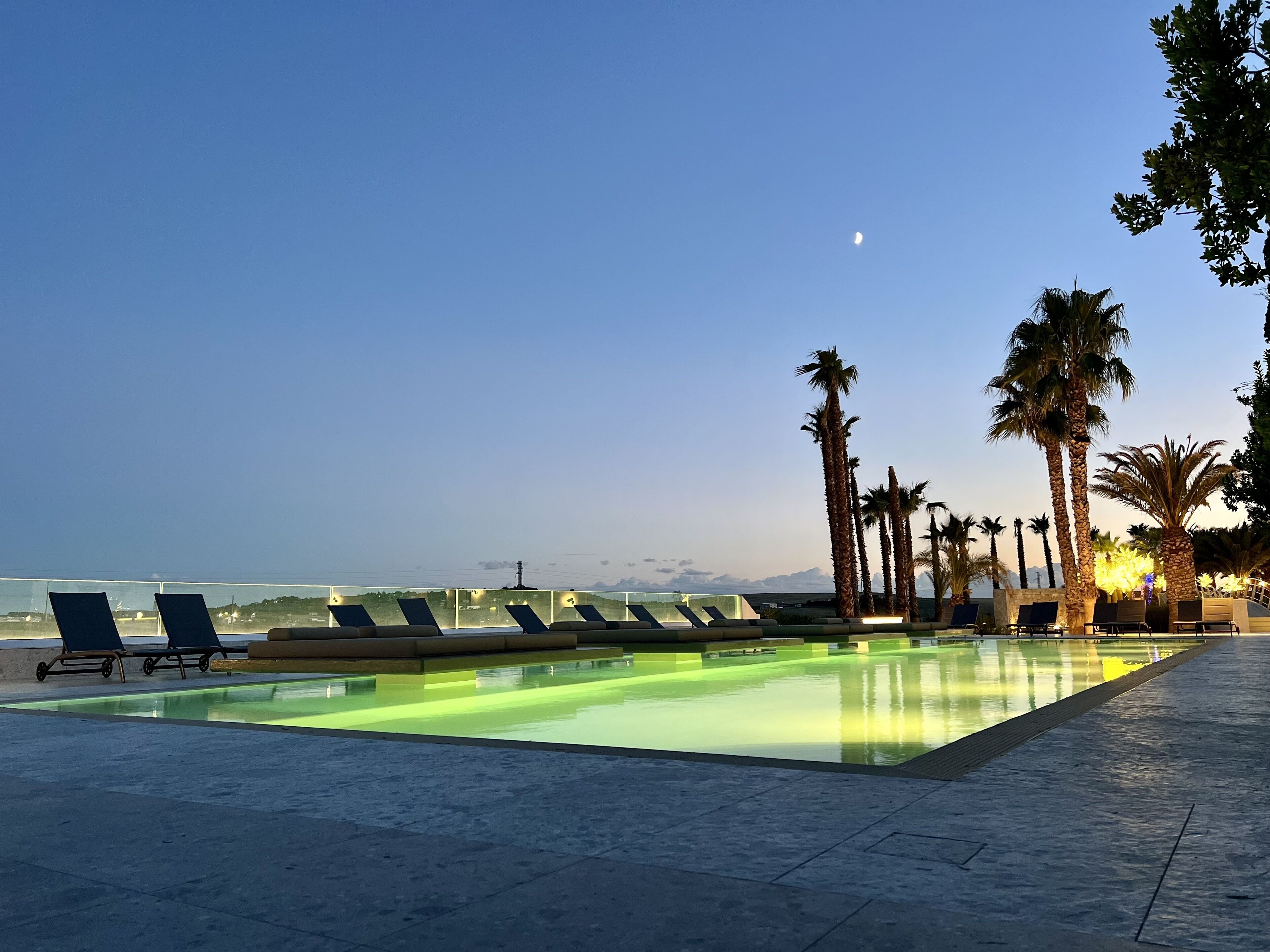 A poolside area with lounge chairs and palm trees, a clear sky, and a distant view of mountains.