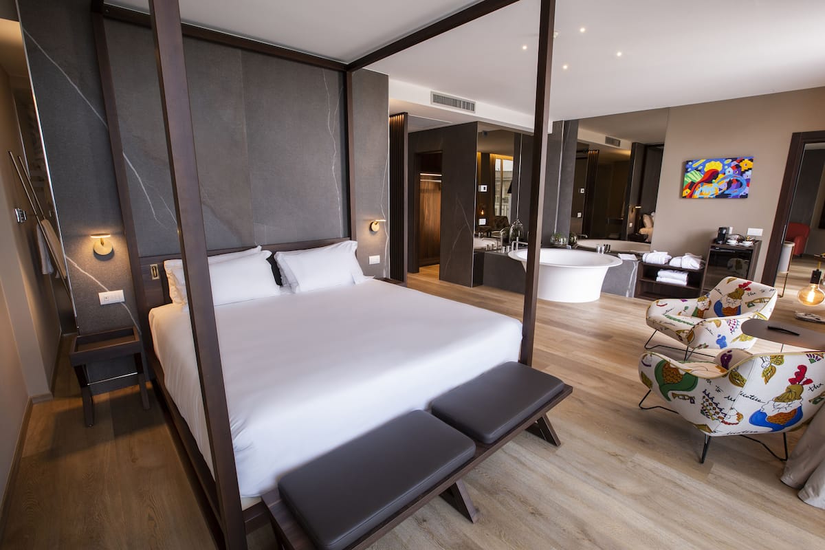 A modern hotel room with a large bed, bench, and a bathroom with a bathtub and shower.