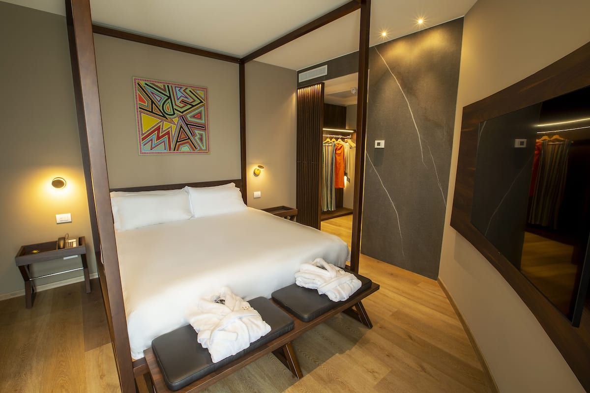 A modern hotel room with a large bed, a bedside table, a bench with towels, a mirror, and a colorful abstract painting.