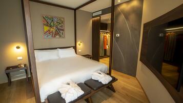 Una camera d'albergo moderna con un letto grande, un comodino, una panca con asciugamani, uno specchio e un quadro astratto dai colori vivaci.
