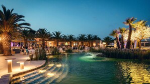 A well-lit outdoor area with a pool, palm trees, and a building with multiple seating areas. - UNA HOTELS MH Matera (Borgo Venusio)