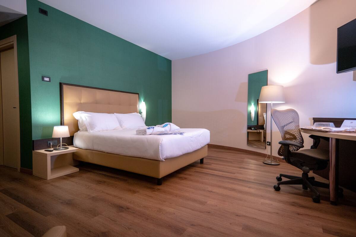 A hotel room with a bed, bedside table, desk, chair, and a TV on the wall.