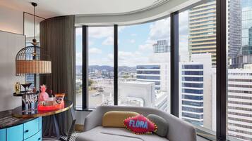 A modern hotel room with a large window offering a city view, a curved desk with a decorative lamp, and a comfortable seating area with a pillow labeled "FLORA".