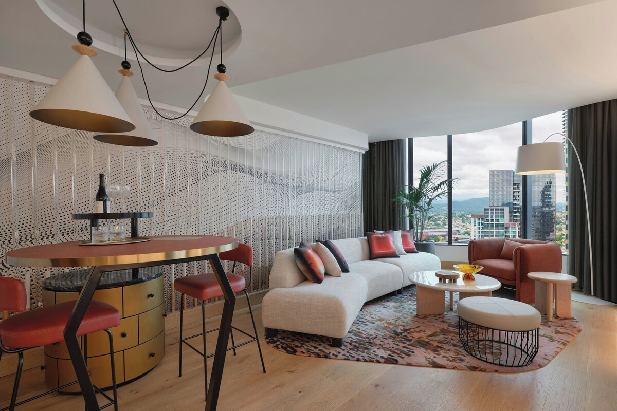 A modern living room with a sofa, coffee table, and chairs. There is a dining table with stools. The room has large windows with a city view.