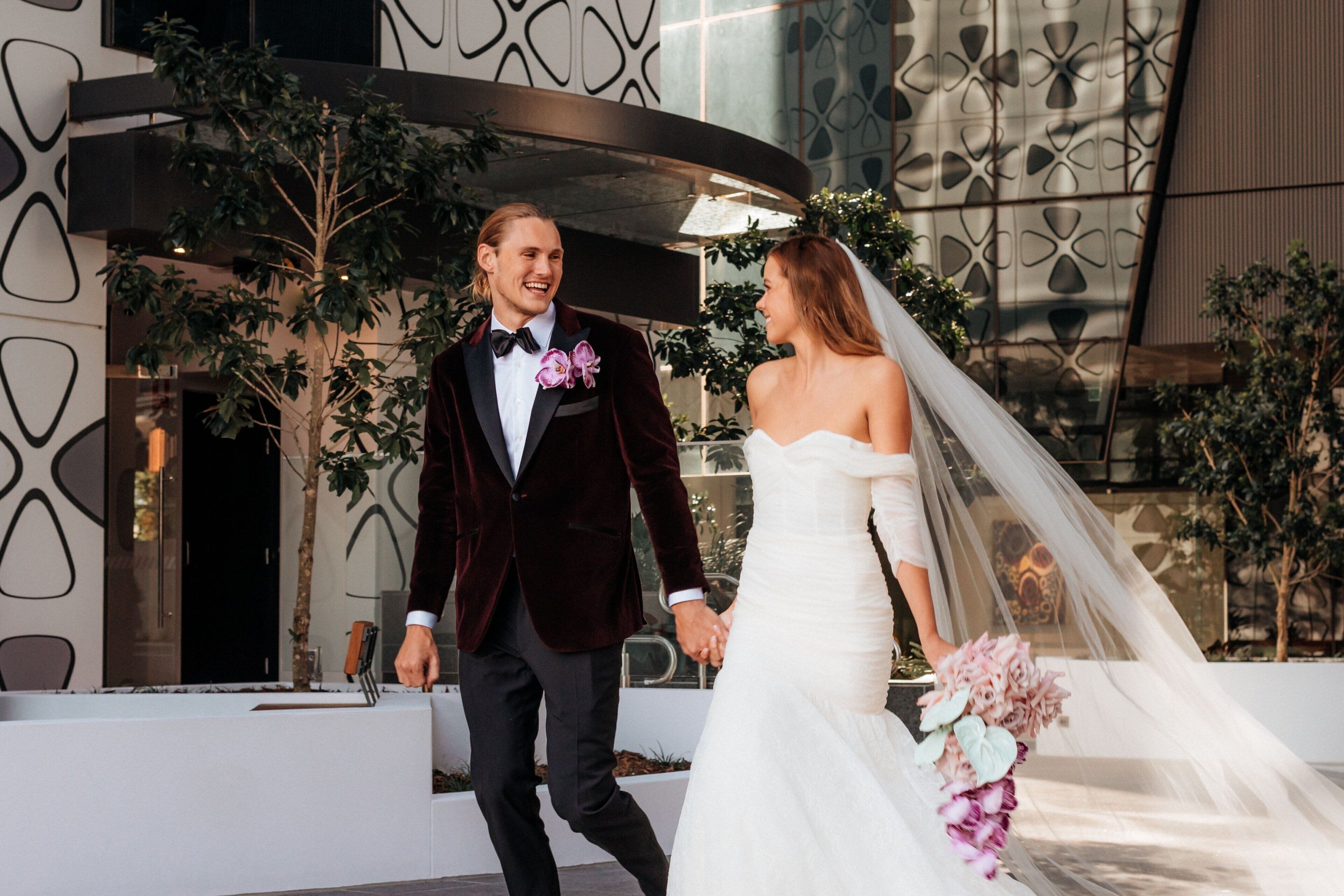 A couple walking hand in hand in front of a modern building with geometric patterns and indoor plants.