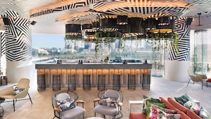 A modern bar area with a city view, featuring geometric ceiling patterns, hanging plants, and contemporary seating.