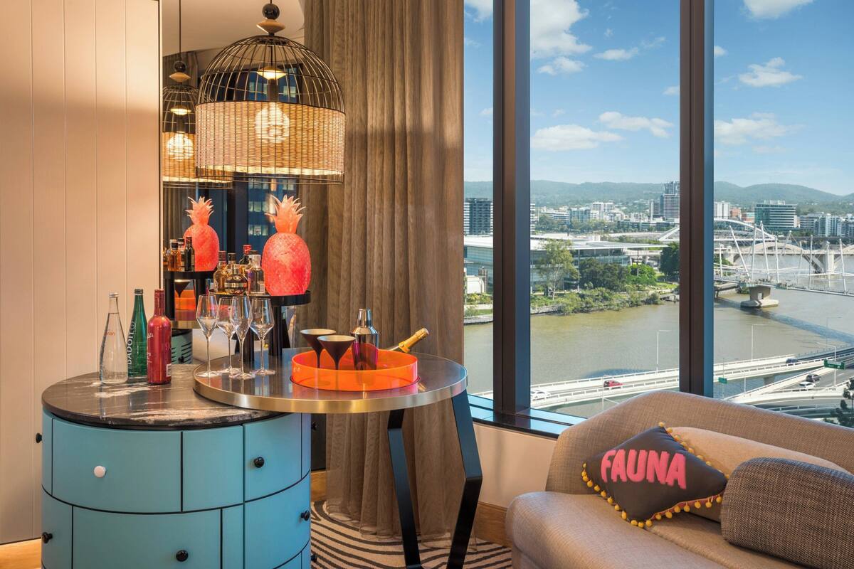 Un salon moderne avec une table ronde sur laquelle sont disposées des boissons, un canapé agrémenté d’un coussin et une vue sur un paysage urbain et une rivière.