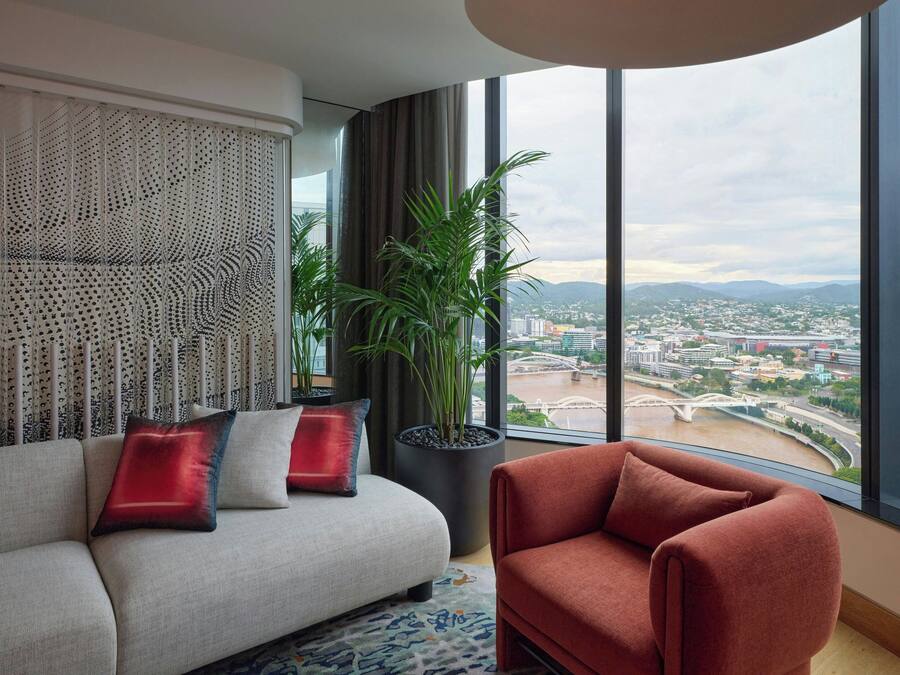 Ein modernes Wohnzimmer mit einer Couch, einem roten Sessel und einer großen Topfpflanze. Eine dekorative Wandverkleidung und der Blick auf eine Stadtlandschaft und einen Fluss sind durch große Fenster sichtbar.