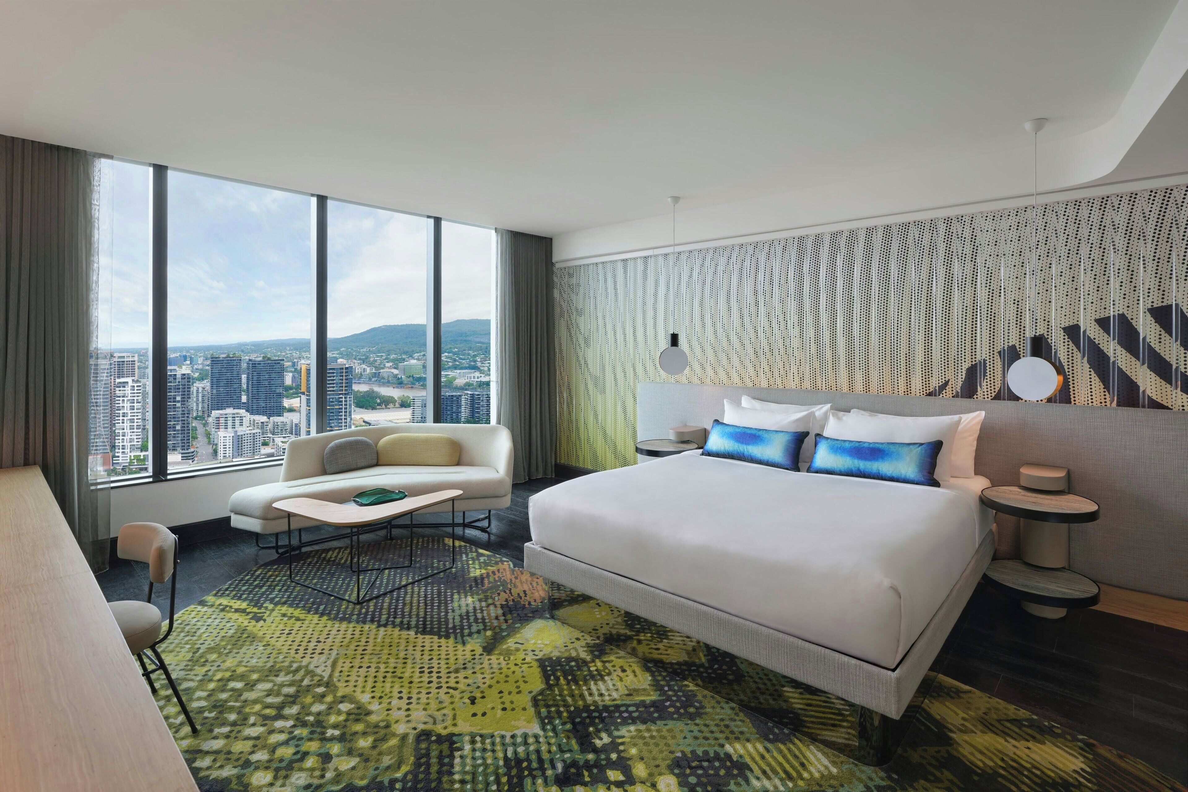A modern hotel room with a large bed, a sofa, a dining table, and a view of the cityscape.