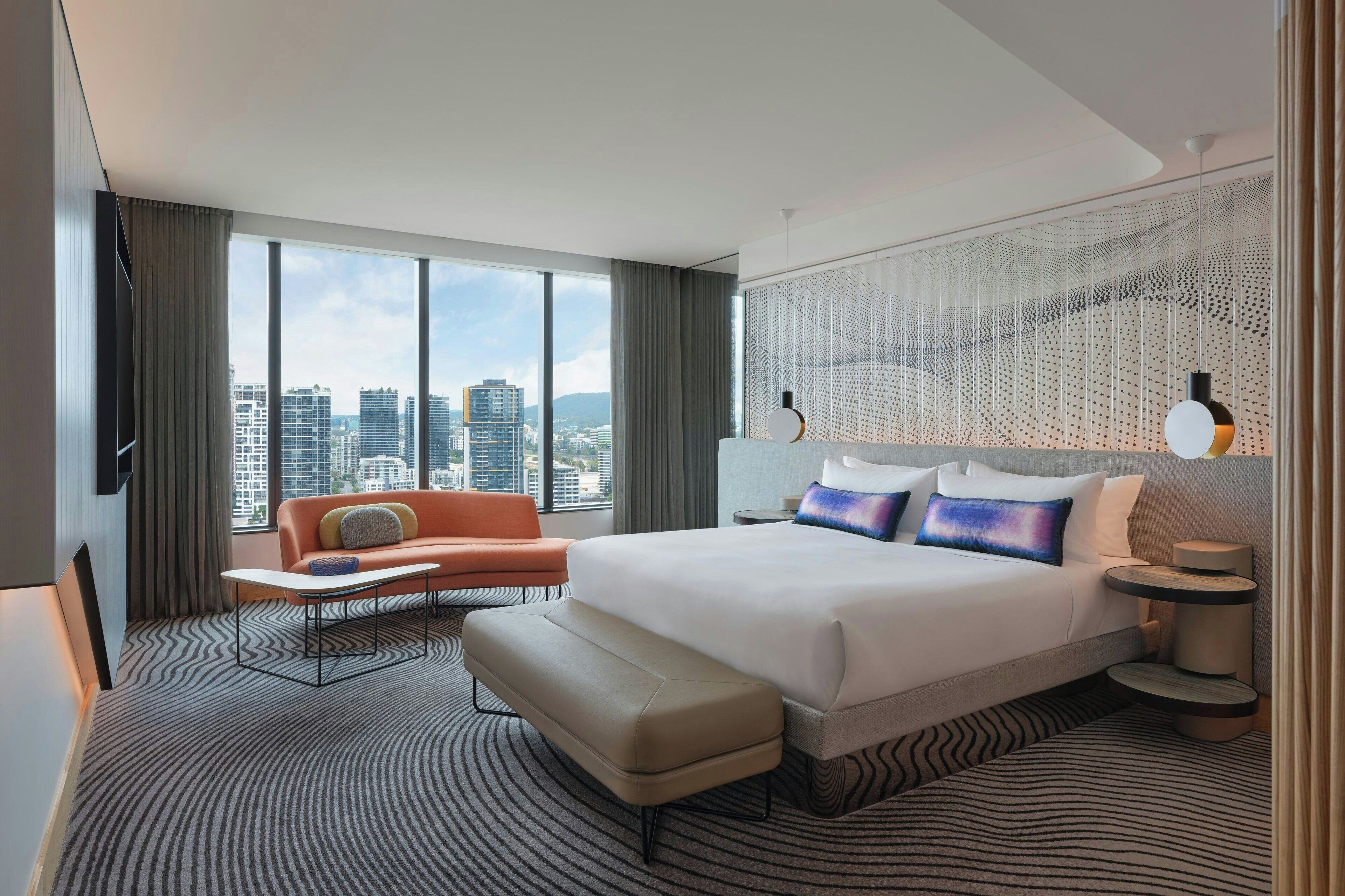 A modern hotel room with a large bed, a sofa, a small table, and a view of the cityscape.
