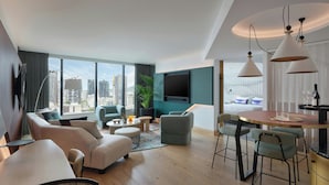 A modern hotel room with a large window, a flat-screen TV, a dining area with a round table, and a seating area with sofas and chairs.