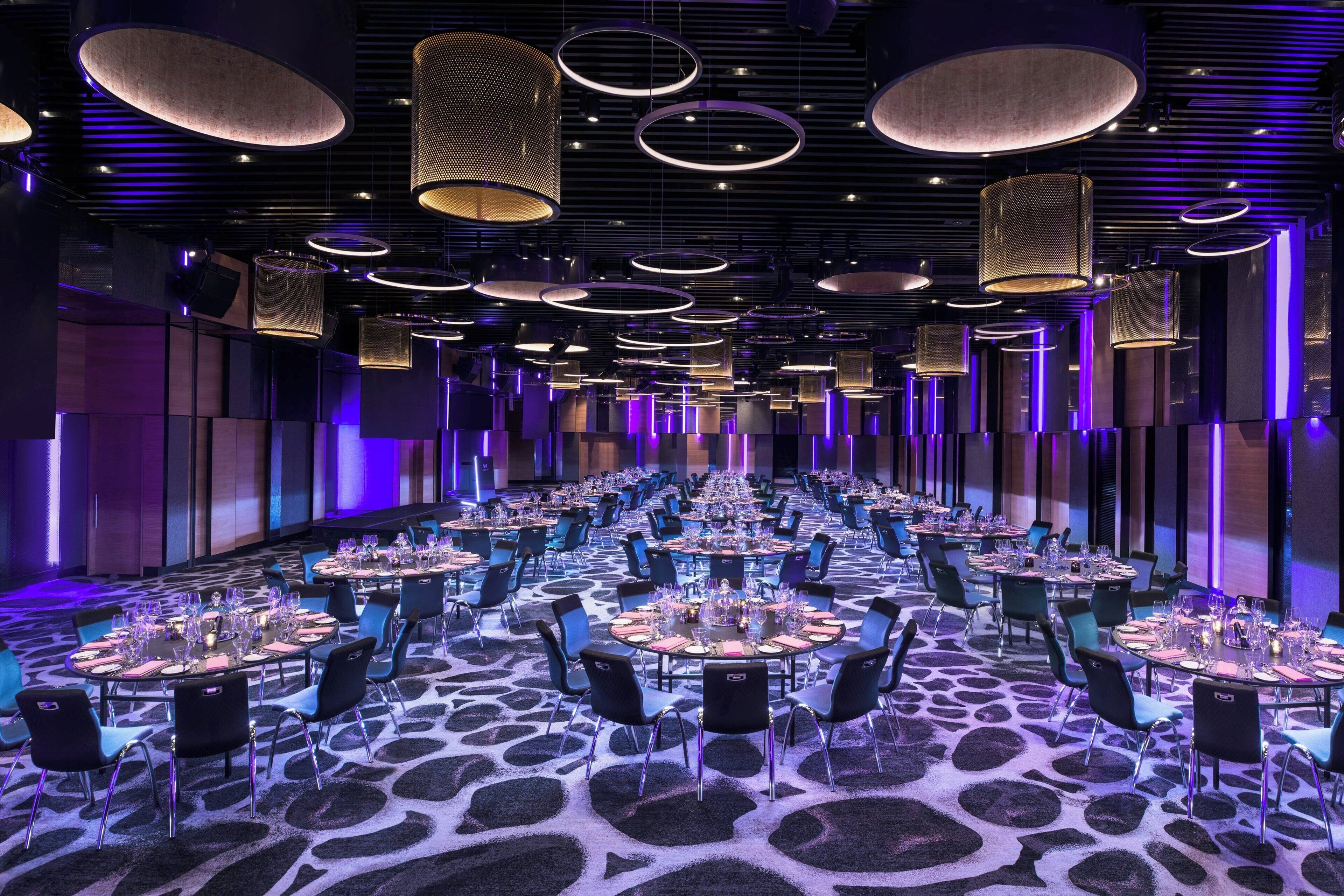 A large banquet hall with round tables set for dining, featuring a patterned carpet and modern lighting fixtures.
