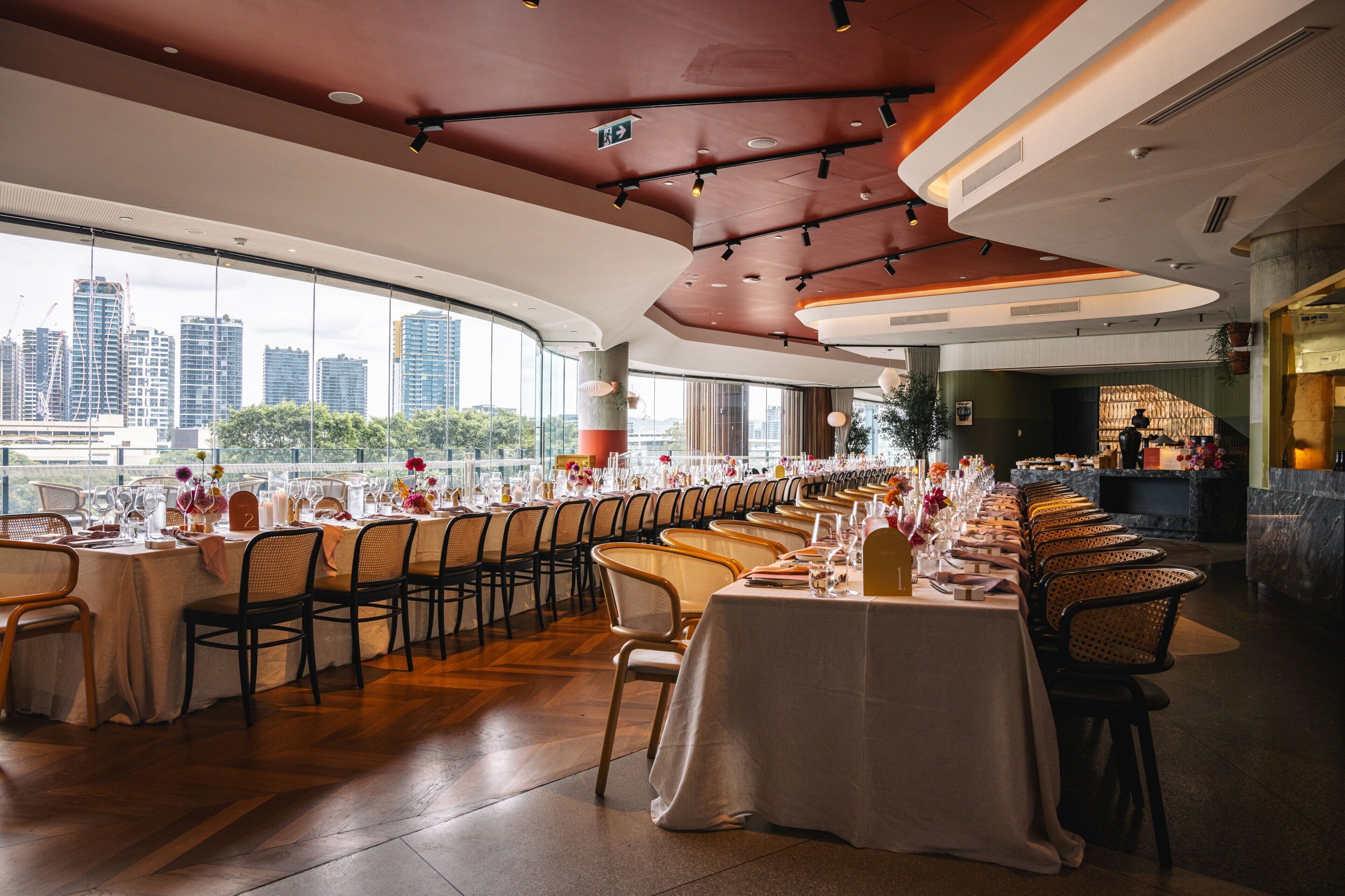 A long dining table set for a formal event with a city view through large windows.