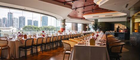 A long dining table set for a formal event with a city view through large windows.