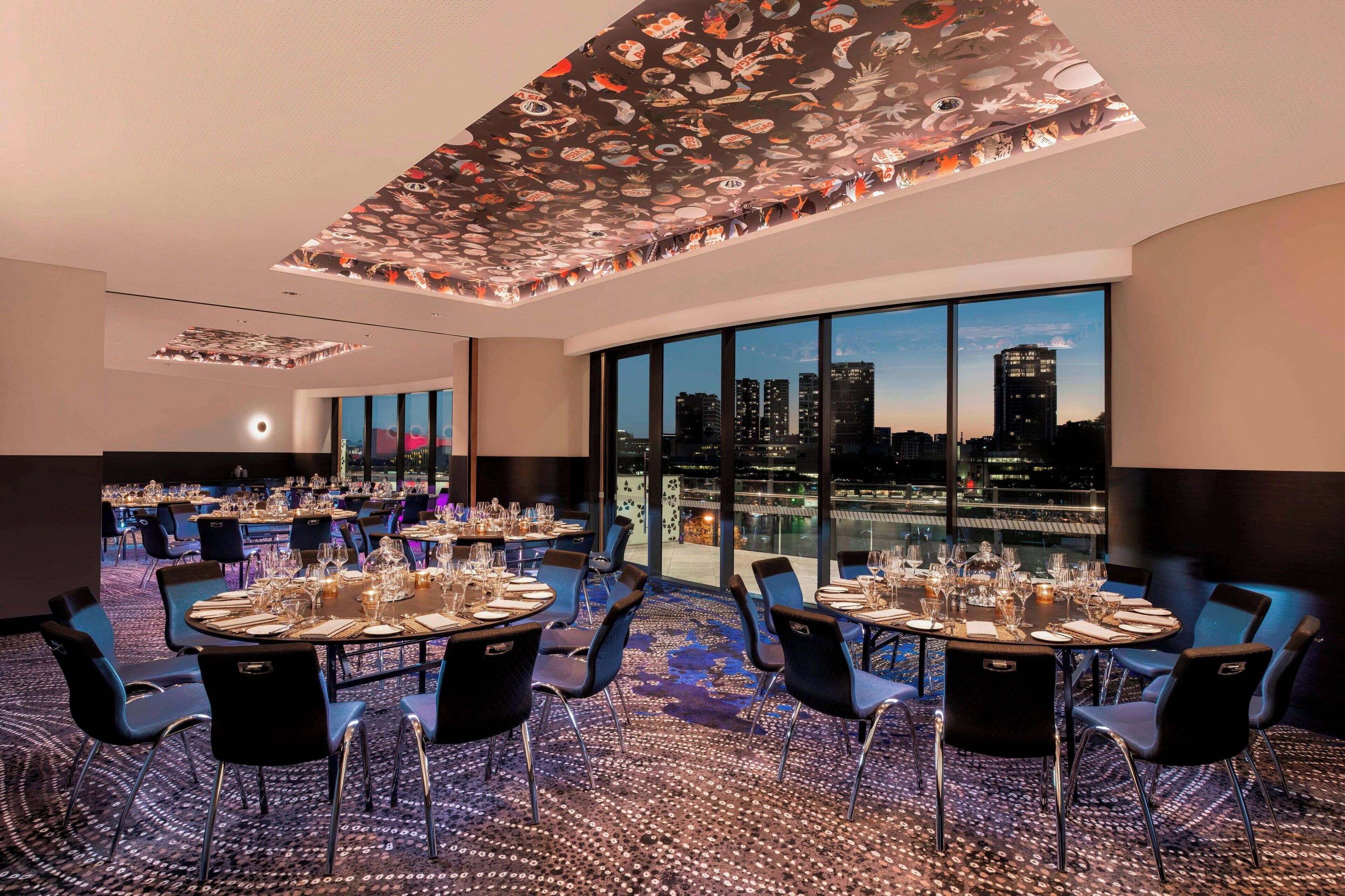 A dining area with round tables set for six, large windows offering a city view, and a patterned ceiling.