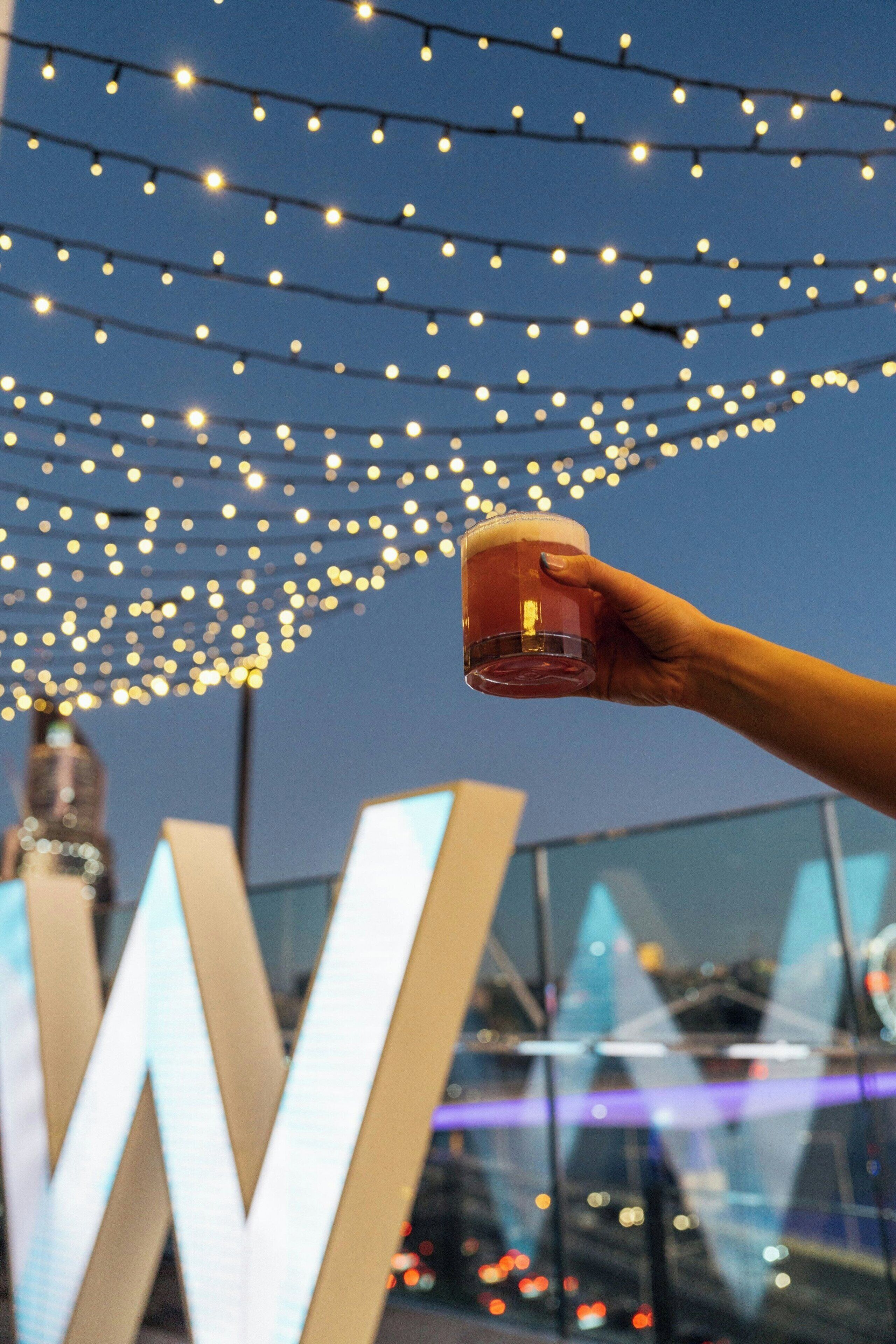 A hand holding a glass of beer with string lights in the background.