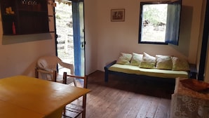 Une pièce avec un plancher en bois, une table jaune, un canapé avec des coussins à motifs, une chaise, une étagère avec des bouteilles et une fenêtre donnant sur l’extérieur.
