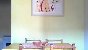 Un coin repas avec une table et des chaises en bois, un tableau abstrait encadré au mur et, au fond, un espace cuisine.
