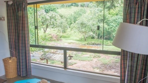 A room with a large window overlooking a rural landscape, a bed, a table with a hat and candles, and a lamp. - Apto 2 Férias na Fazenda Usina Térreo (Bocaina de Minas)