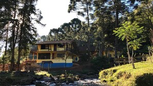 A two-story building with a balcony, surrounded by trees and a flowing stream. - Apto 2 Férias na Fazenda Usina Térreo (Bocaina de Minas)