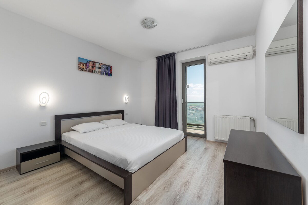 A bedroom with a bed, bedside tables, a dresser, a window with curtains, an air conditioning unit, and a cityscape view.