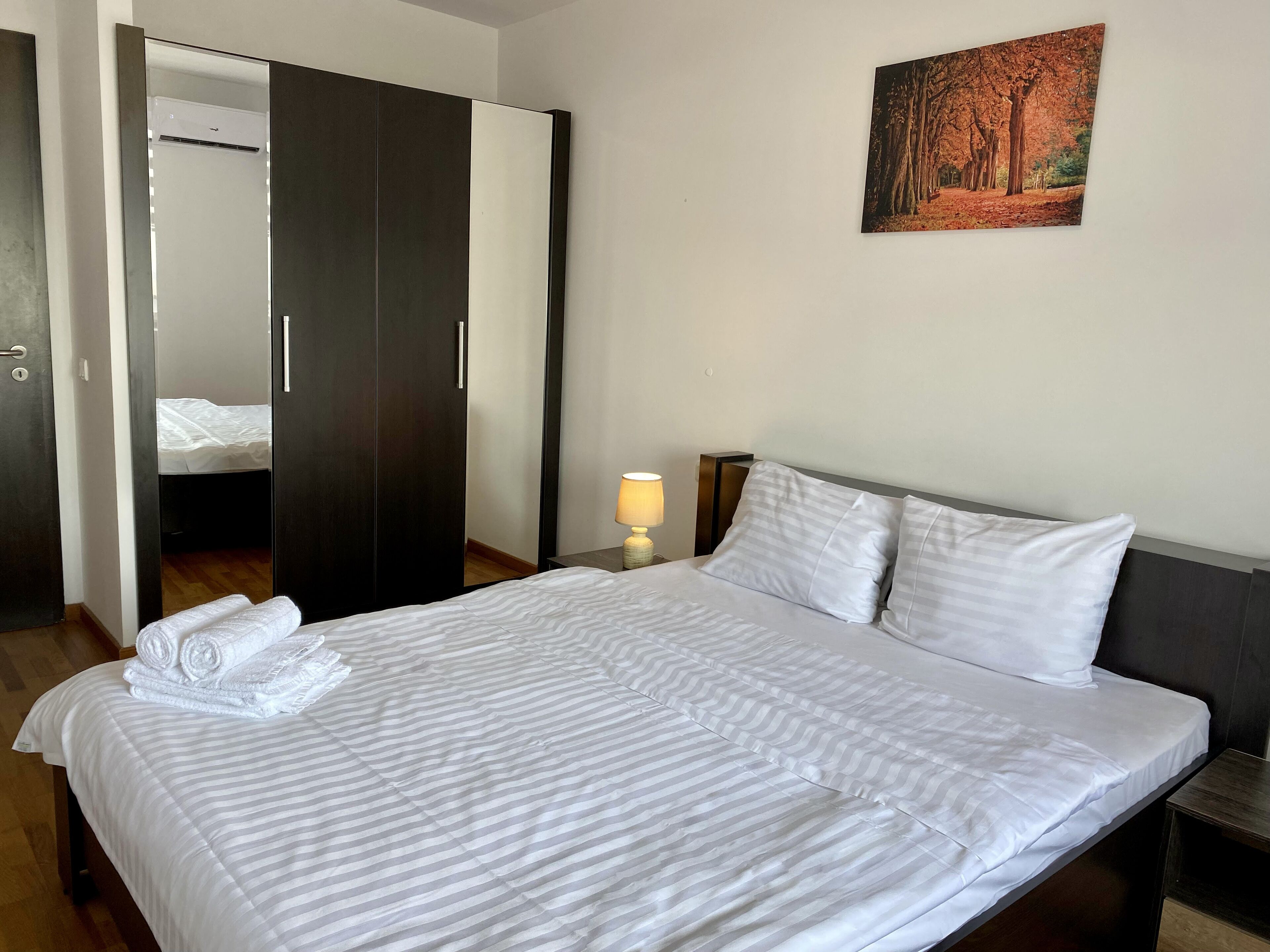 A neatly made bed with white bedding, a wooden wardrobe, a bedside lamp, and a framed picture on the wall.