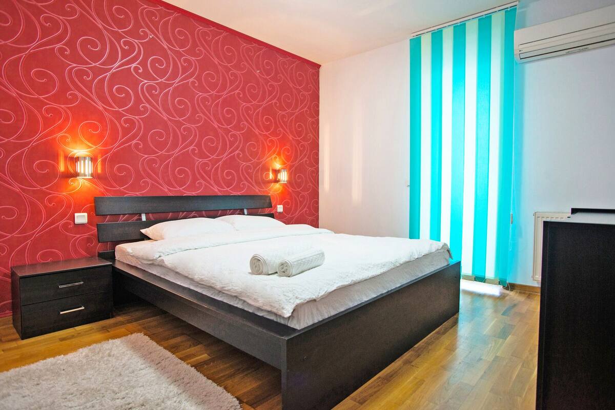 Ett modernt sovrum med en röd, mönstrad vägg, två sänglampor, en säng med vita sängkläder, ett trägolv och utsikt över en blå- och vitrandig gardin.