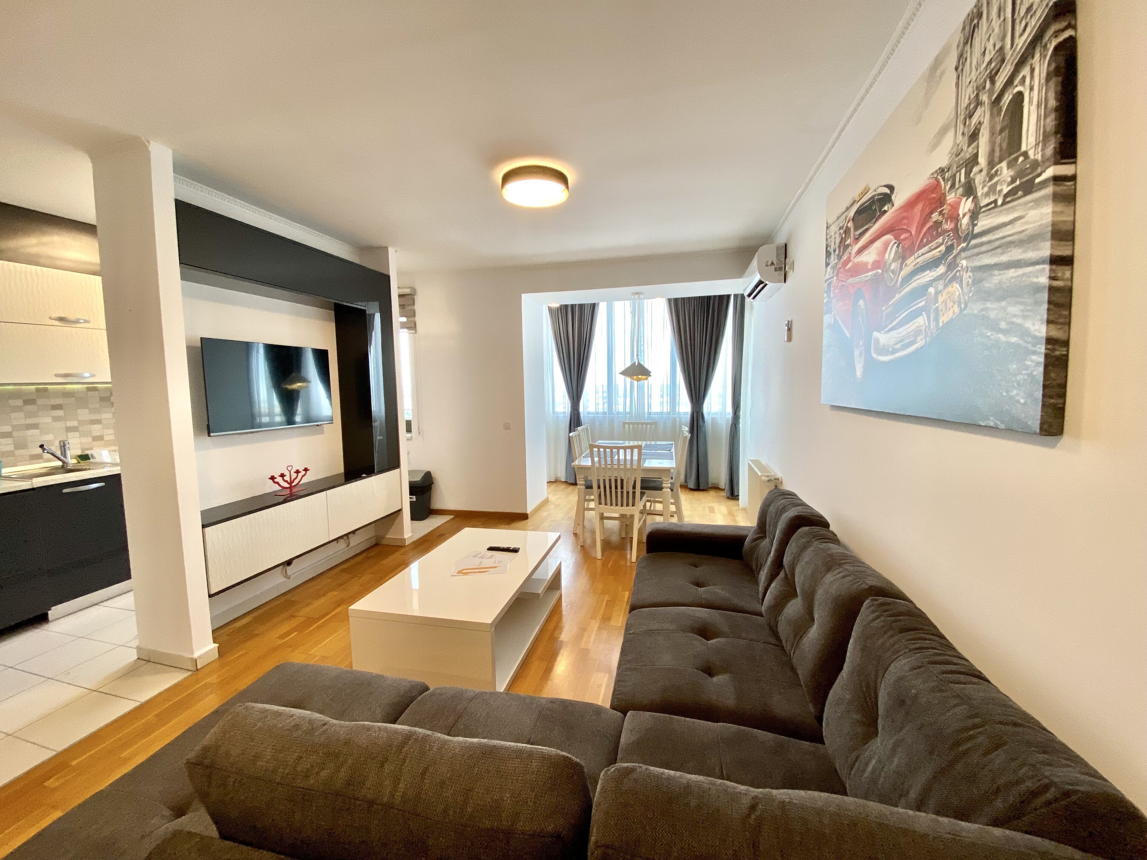 A modern living room with a sofa, a dining area with a table and chairs, a kitchen area with a TV, and a painting on the wall.