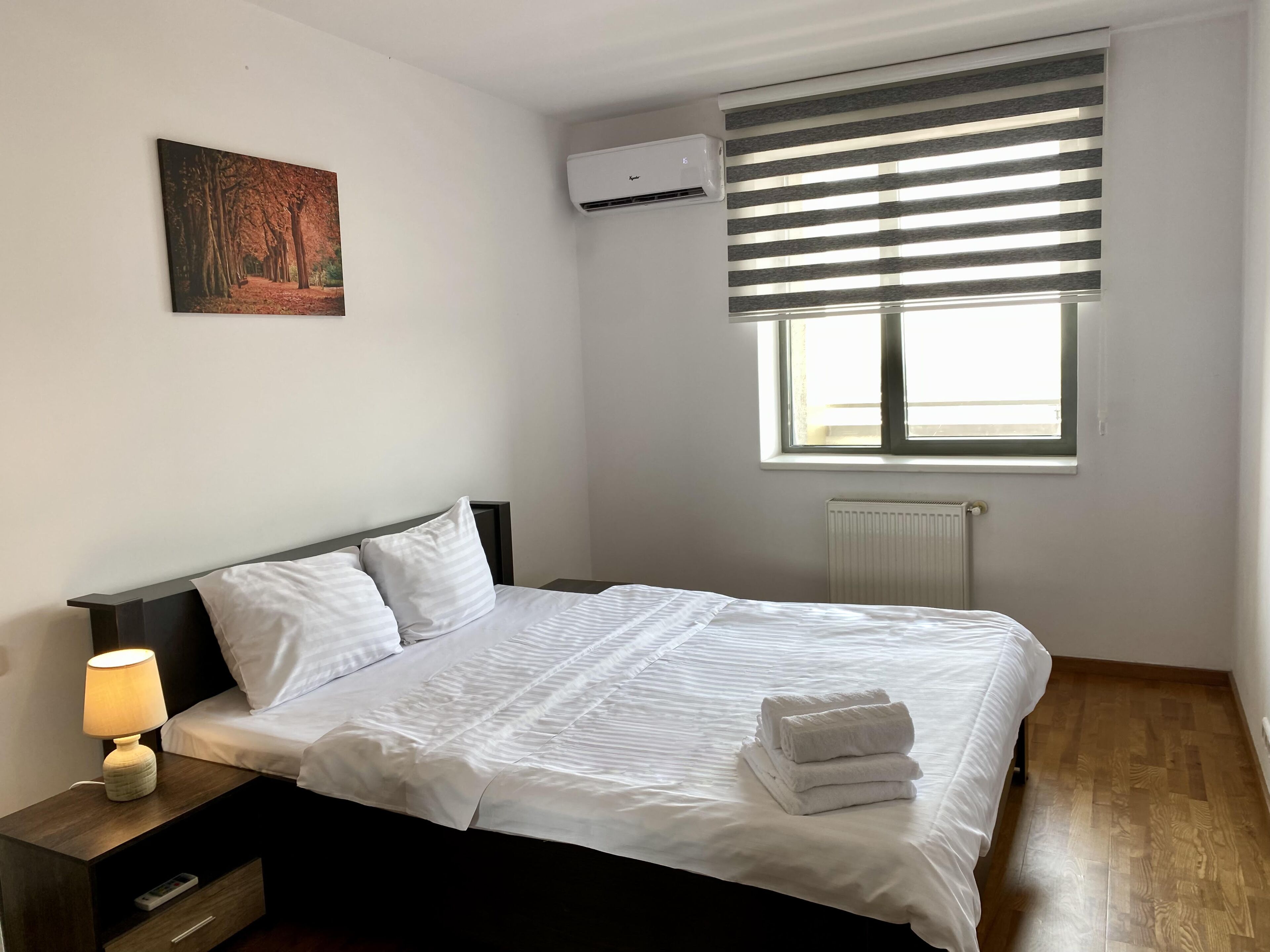 A neatly made bed with white linens and a stack of towels, a bedside lamp, a nightstand with a remote control drawer, a radiator, and a window with blinds.