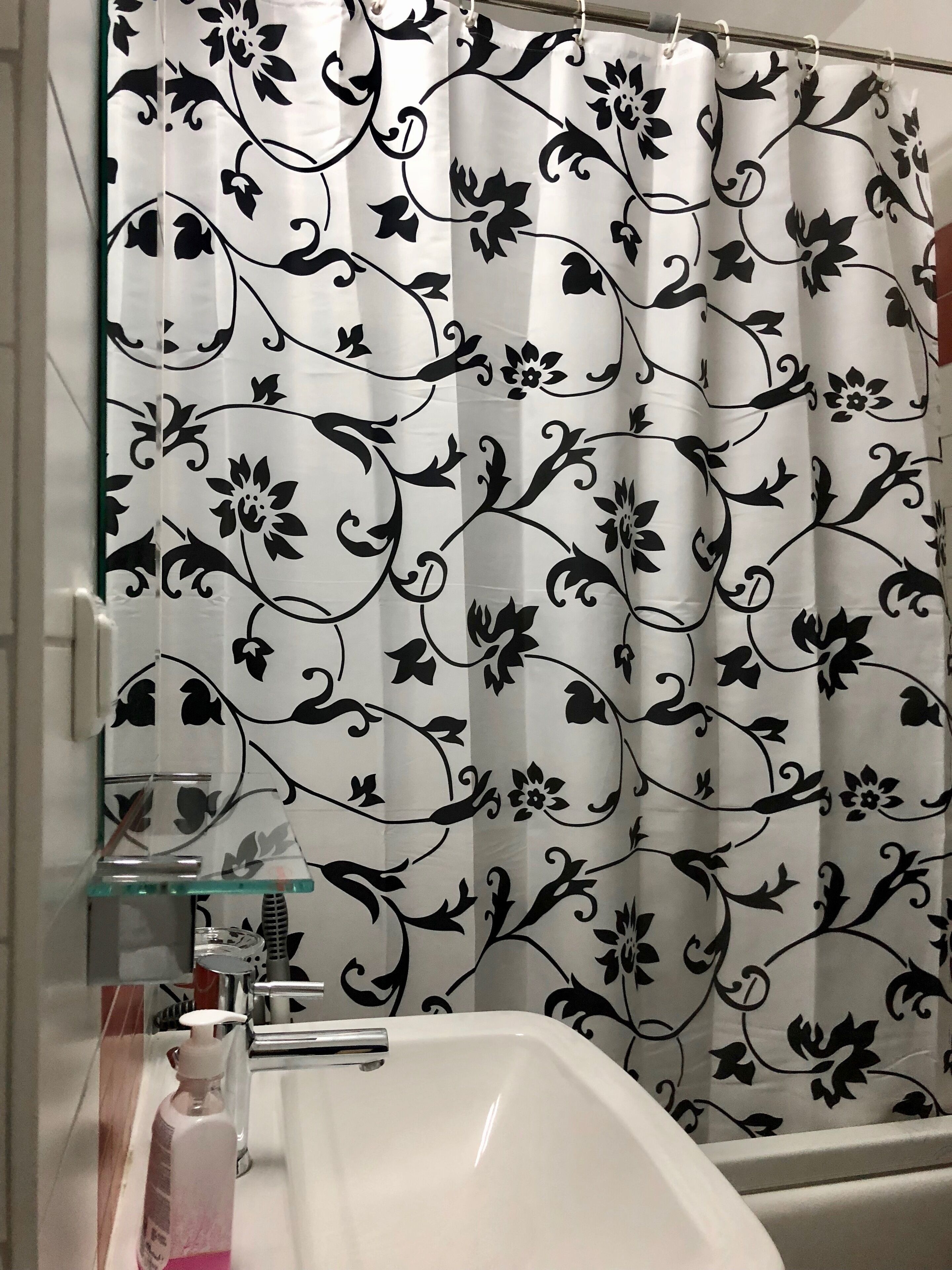 Une salle de bain avec un rideau de douche orné d’un motif floral noir, une étagère en verre où sont posés des articles de toilette et un lavabo blanc.