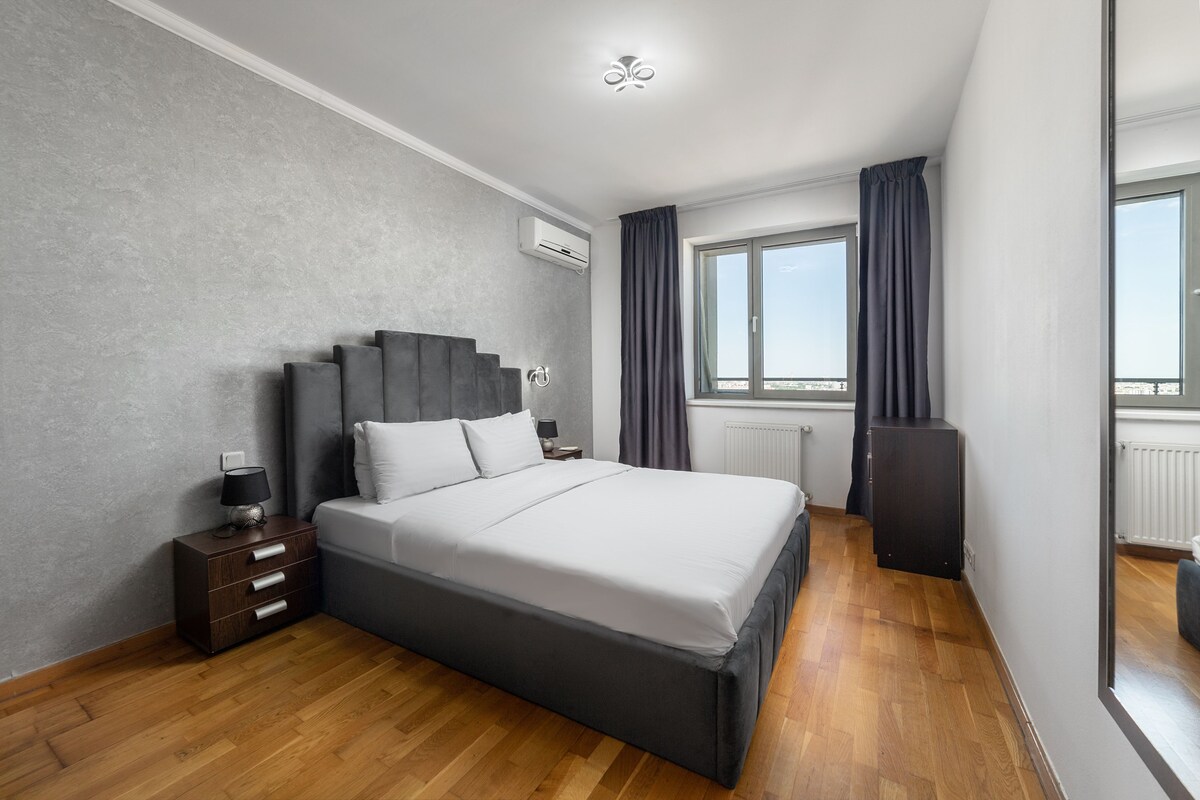A modern bedroom with a large bed, a nightstand, a dresser, and a window with curtains.