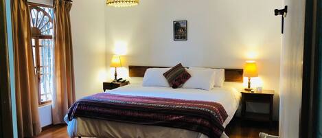 A neatly arranged hotel room with a large bed, two bedside lamps, a hanging light fixture, a small bedside table, and a radiator.