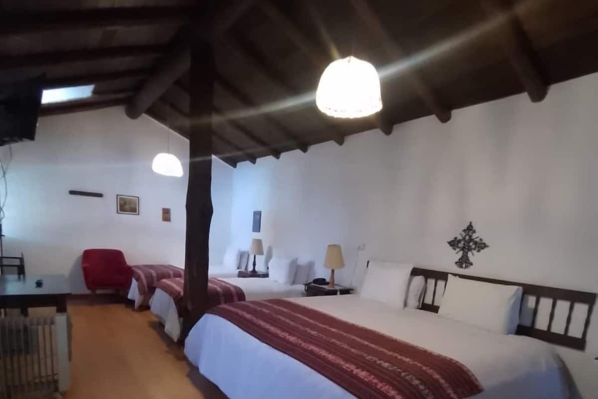 Una habitación con piso de madera, una cama con cubrecama rojo y blanco, una silla roja, un escritorio con lámpara y una cruz decorativa colgada en la pared.