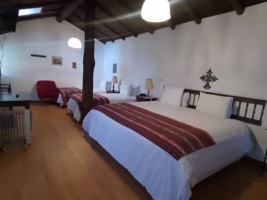 Une pièce avec un plancher en bois, un lit recouvert d’une couverture rouge et blanche, une chaise rouge, un bureau avec une lampe et un crucifix fixé au mur.