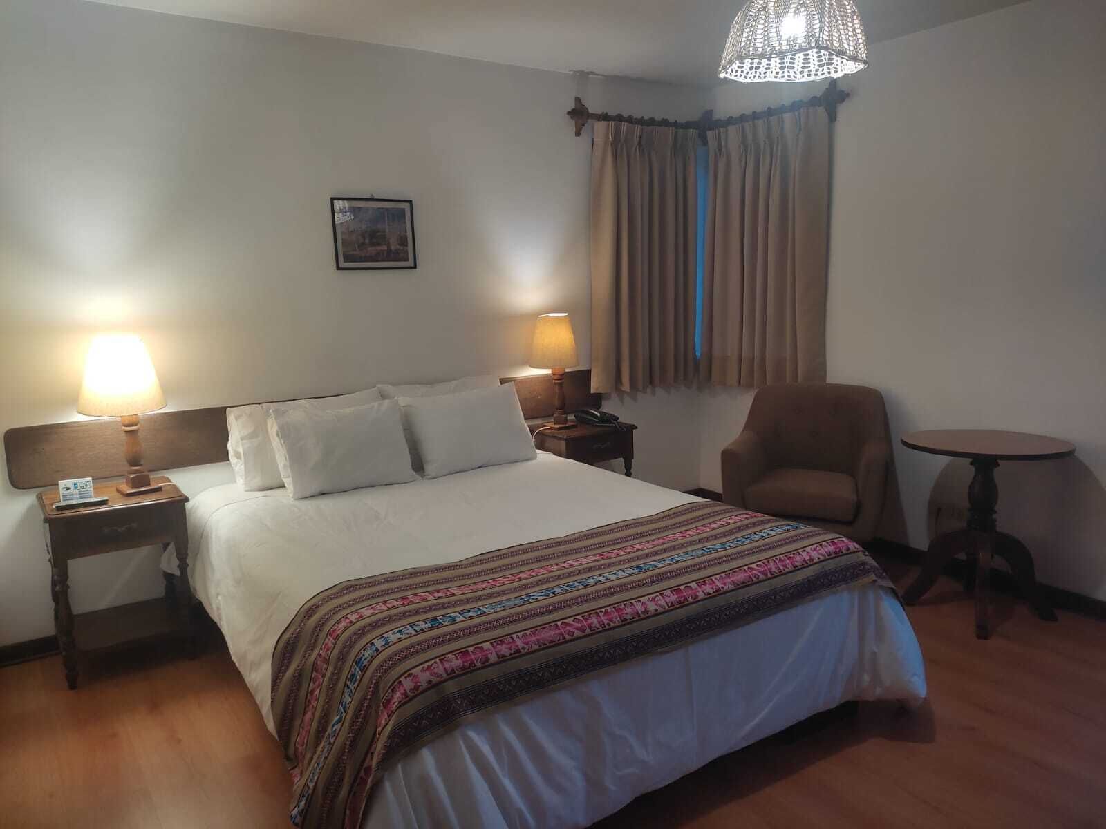 A hotel room with a large bed, two bedside lamps, a chair, a small table, and a window with curtains.