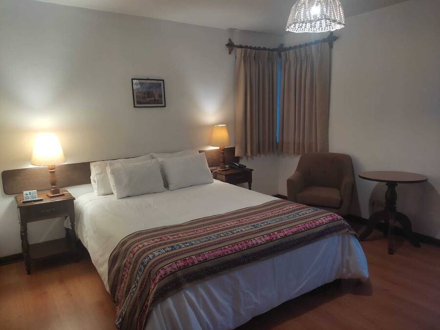 A hotel room with a large bed, two bedside lamps, a chair, a small table, and a window with curtains.