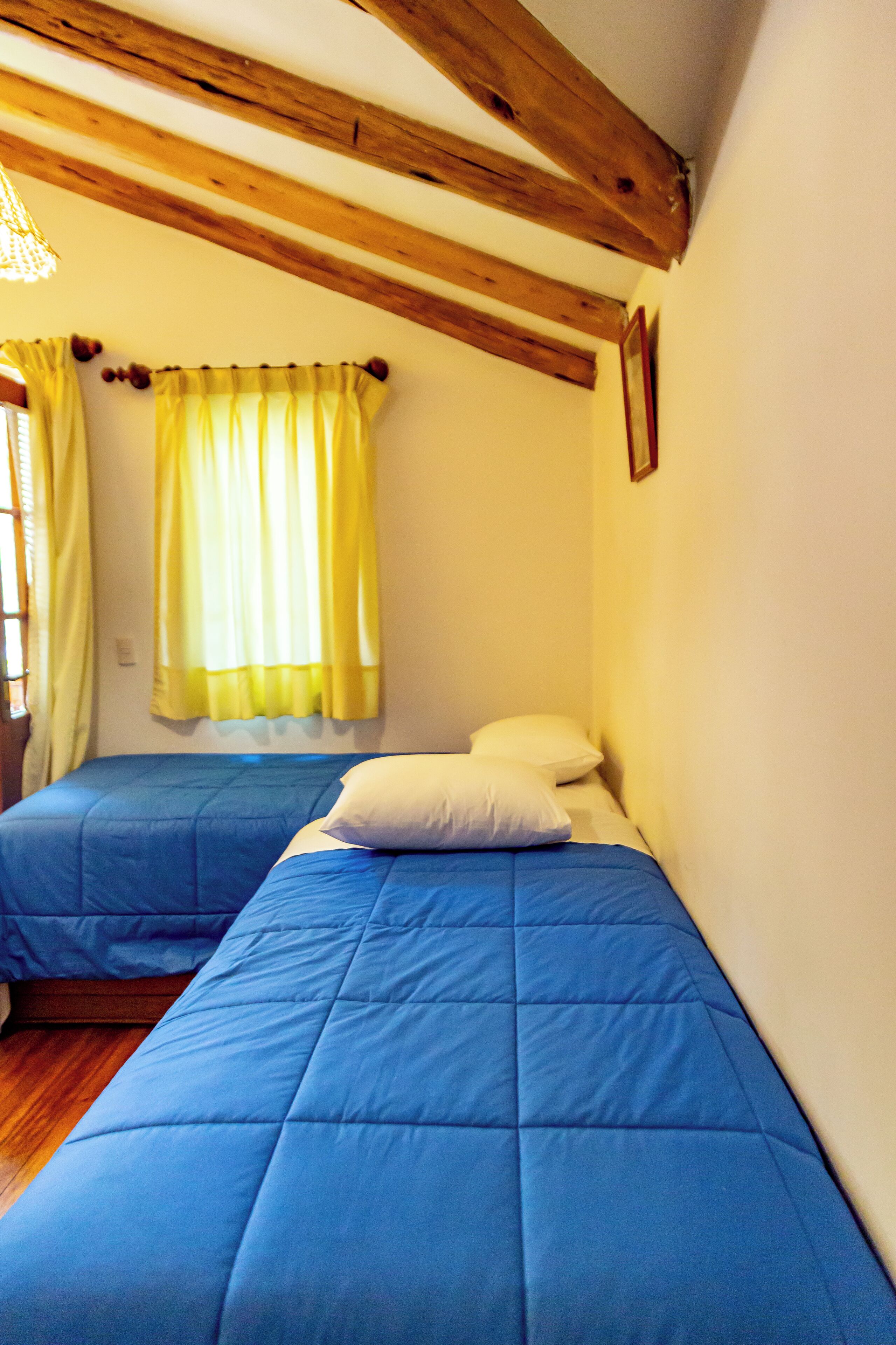 Een kamer met twee bedden, houten balken in het plafond en een raam met gele gordijnen.