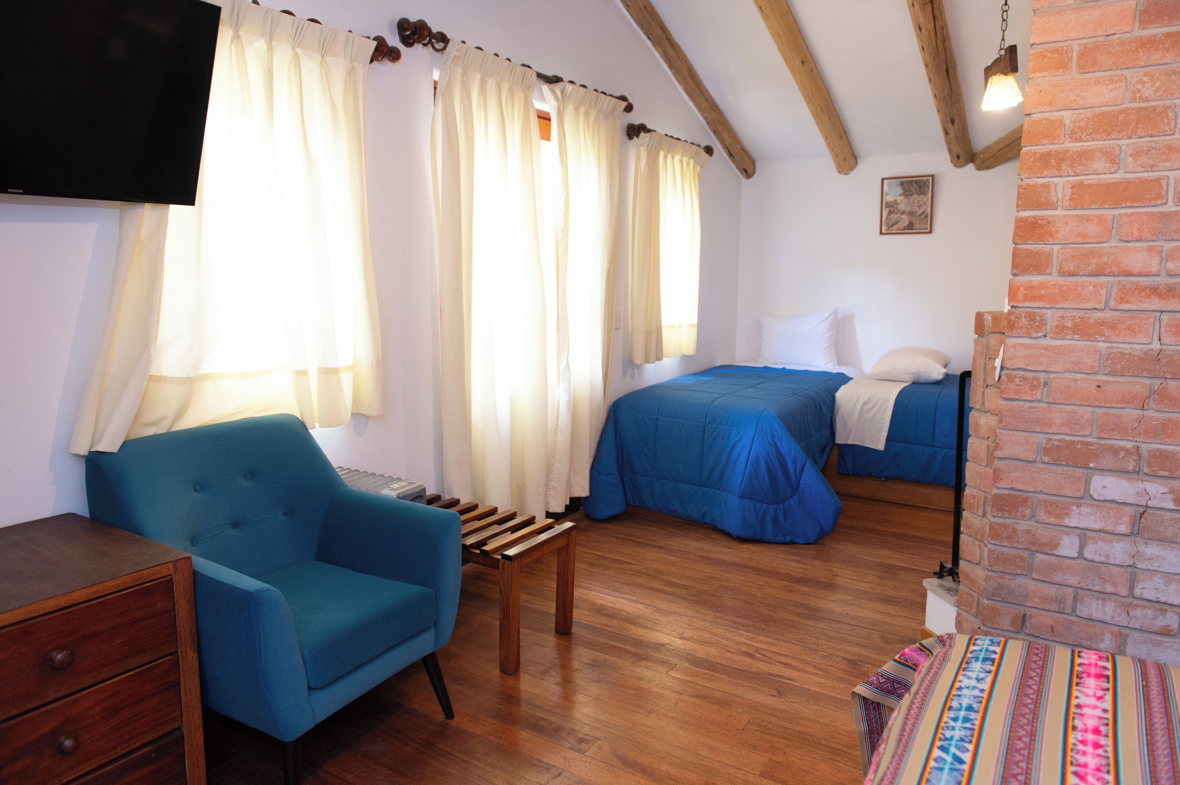 A bedroom with two beds, a blue armchair, a wooden table, and a brick wall.