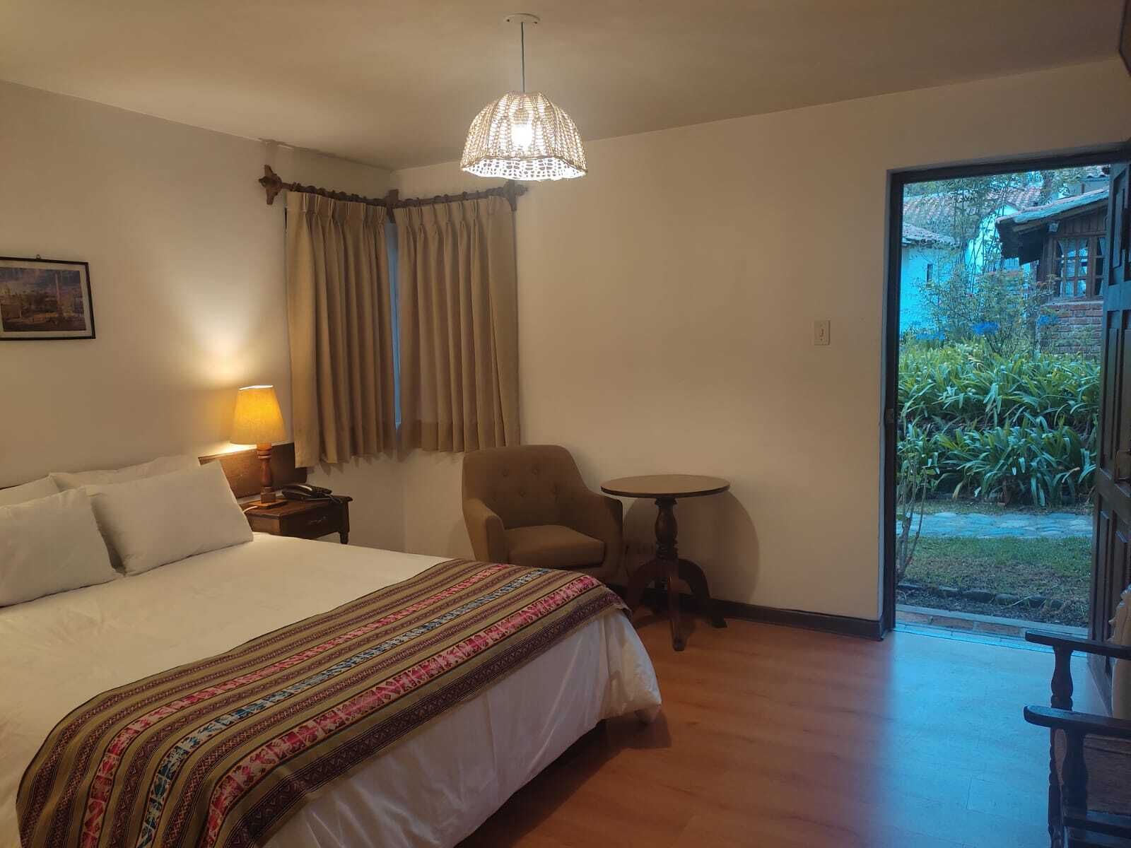 A hotel room with a large bed, a small round table, a chair, a lamp, and a view of the garden through a glass door.