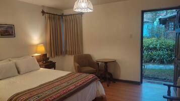 A hotel room with a large bed, a small round table, a chair, a lamp, and a view of the garden through a glass door.