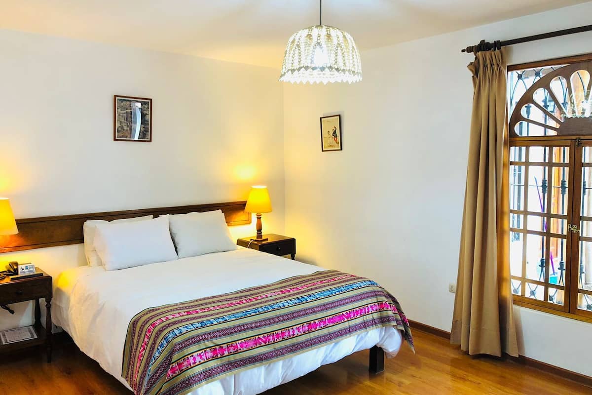 Habitación de hotel con cama, dos lámparas de noche, una lámpara decorativa en el techo, una ventana con cortinas y dos cuadros en la pared.