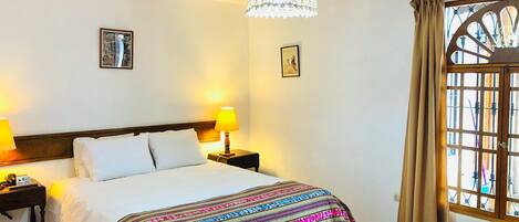Habitación de hotel con cama, dos lámparas de noche, una lámpara decorativa en el techo, una ventana con cortinas y dos cuadros en la pared.