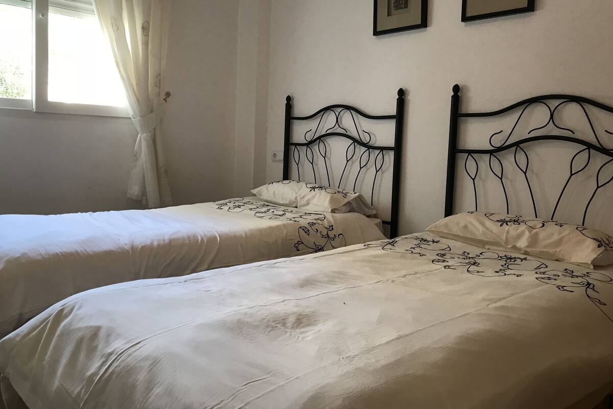 A room with two single beds, each with a metal headboard and white bedding with floral patterns. There are two framed pictures on the wall above the beds.