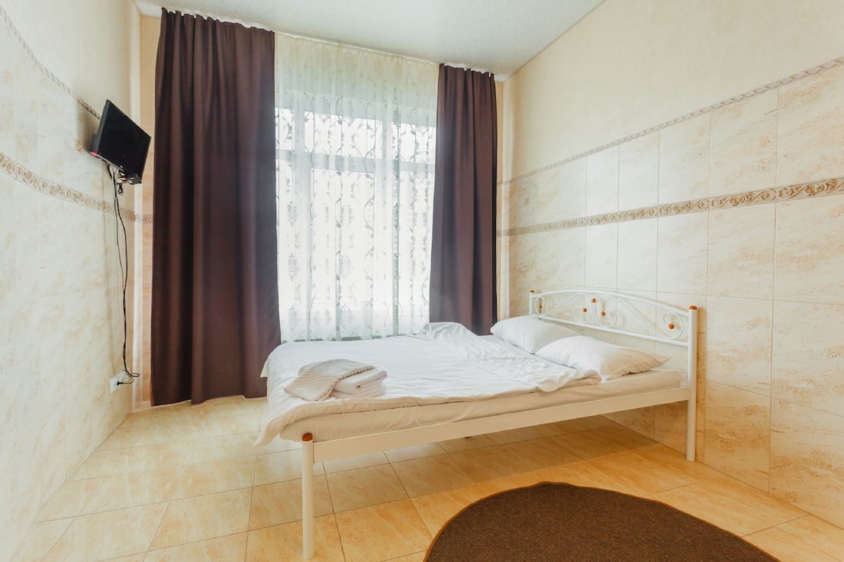 A single-bed room with a white metal frame, a flat-screen TV mounted on the wall, a window with sheer curtains and a dark drape, and a tiled floor.