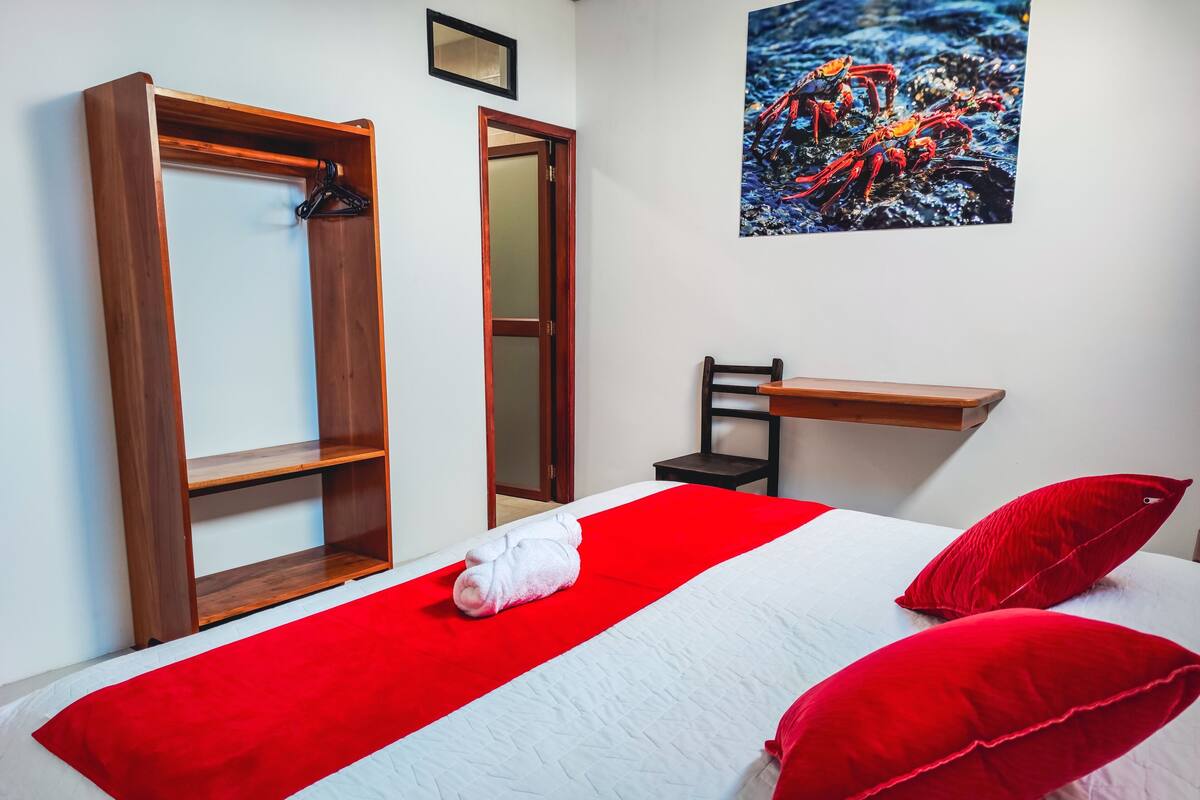 Une chambre à coucher avec un lit, une couvre-lit rouge et blanche, un oreiller rouge, une armoire en bois, un miroir et un tableau accroché au mur.