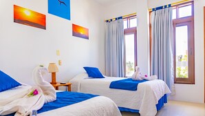 Habitación de hotel con dos camas, cada una con cubrecama azul y blanco, y una toalla con forma de cisne. Hay dos ventanas con cortinas, y un cuadro de un atardecer en la pared.