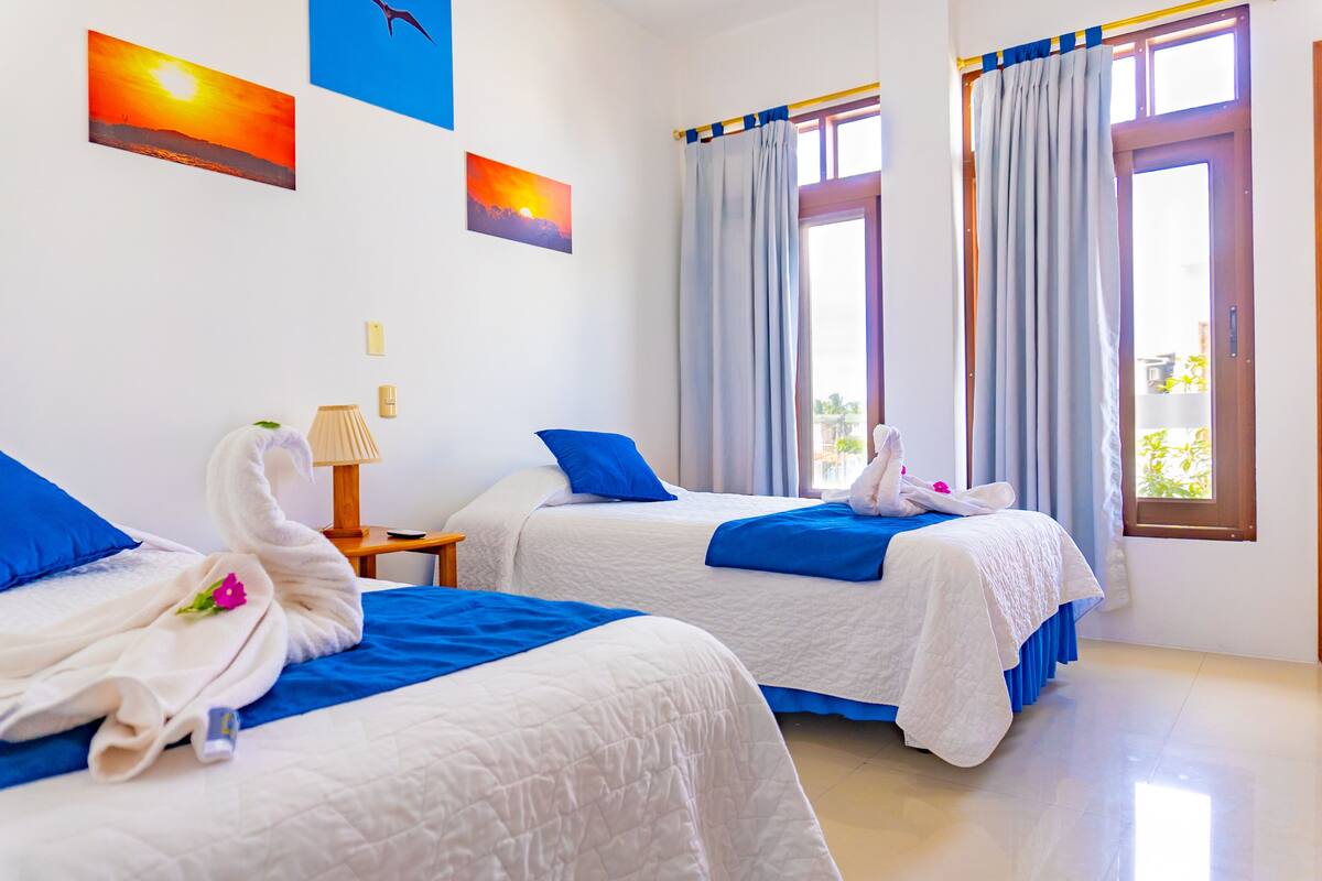 Une chambre d’hôtel avec deux lits, chacun recouvert d’une couette bleue et blanche, et une serviette en forme de cygne. Il y a deux fenêtres avec des rideaux, et un tableau représentant un coucher de soleil est accroché au mur.