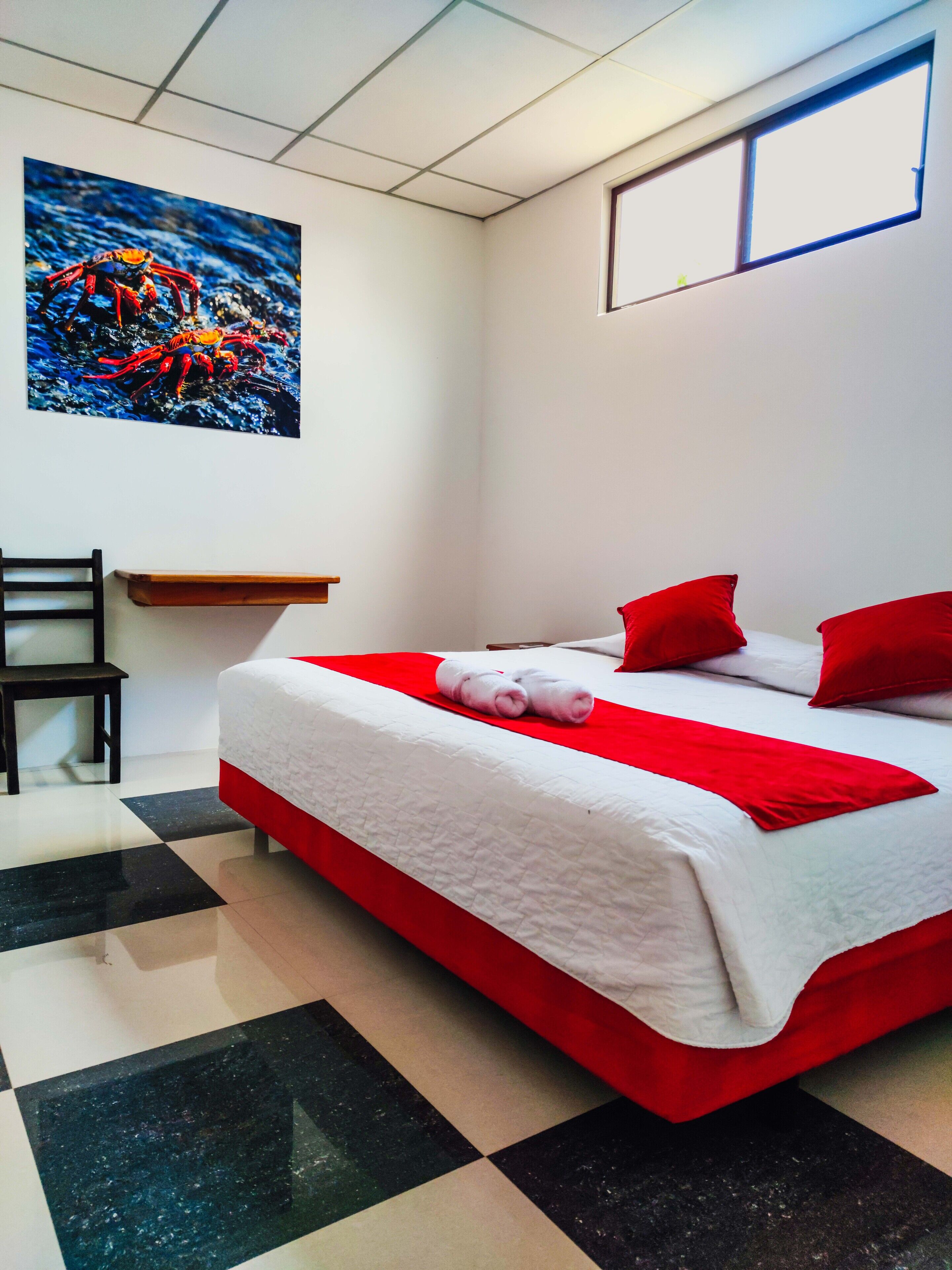 A hotel room with a checkered floor, a bed with red and white bedding, a red pillow, a white pillow, and a painting of a crab on the wall.