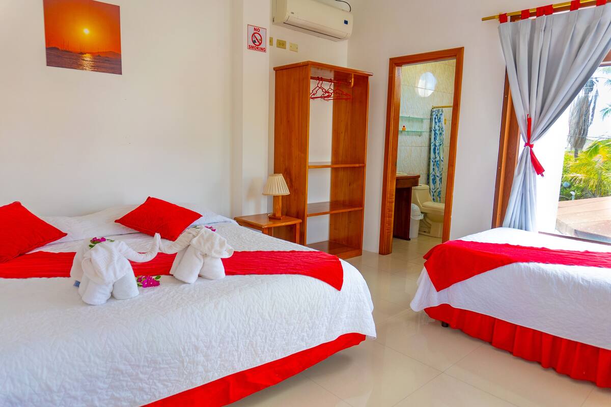 Une chambre d’hôtel avec deux lits, une literie rouge, une armoire en bois, une salle de bain visible à travers une porte ouverte et un tableau représentant un coucher de soleil accroché au mur.